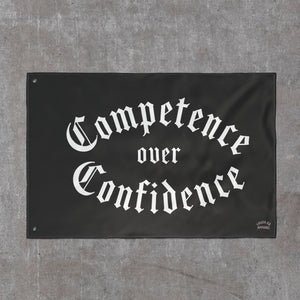 Competence over Confidence flag in white blackletter on black, hung flat against a concrete wall